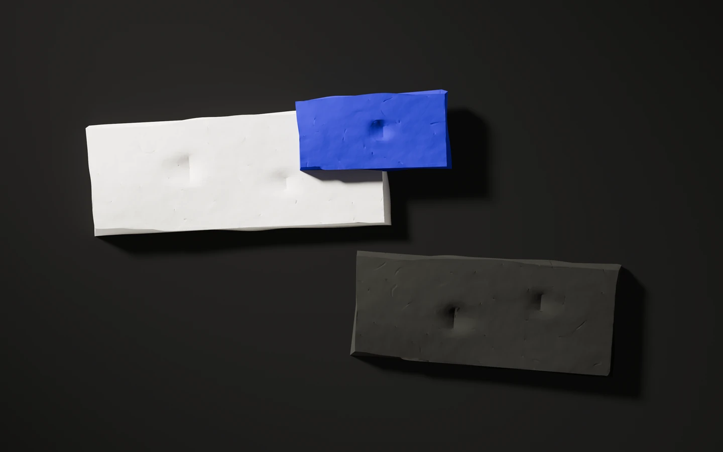 Three matte clay slabs on a dark warm-grey ground, seen from above: one off-white, one blue, one near-black, each pressed with shallow thumbprints and casting a soft shadow towards the lower right.